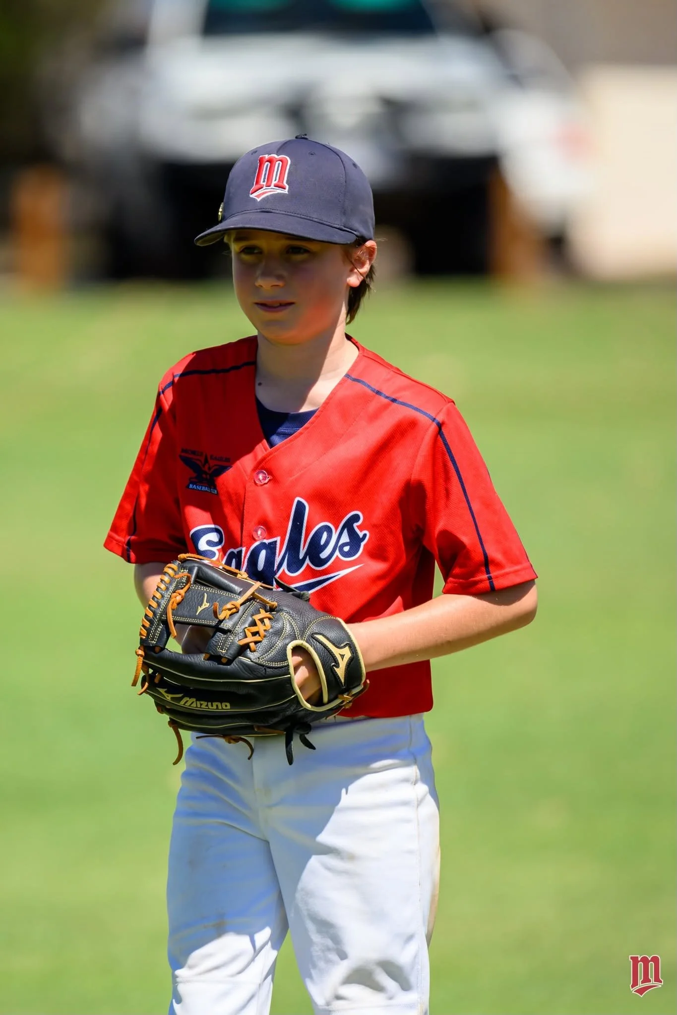 A young baseball player wearing a navy cap with a red and white logo, a red jersey with blue accents, and white pants, standing on a grassy field holding a black and tan baseball glove. Background shows a blurred vehicle and trees.