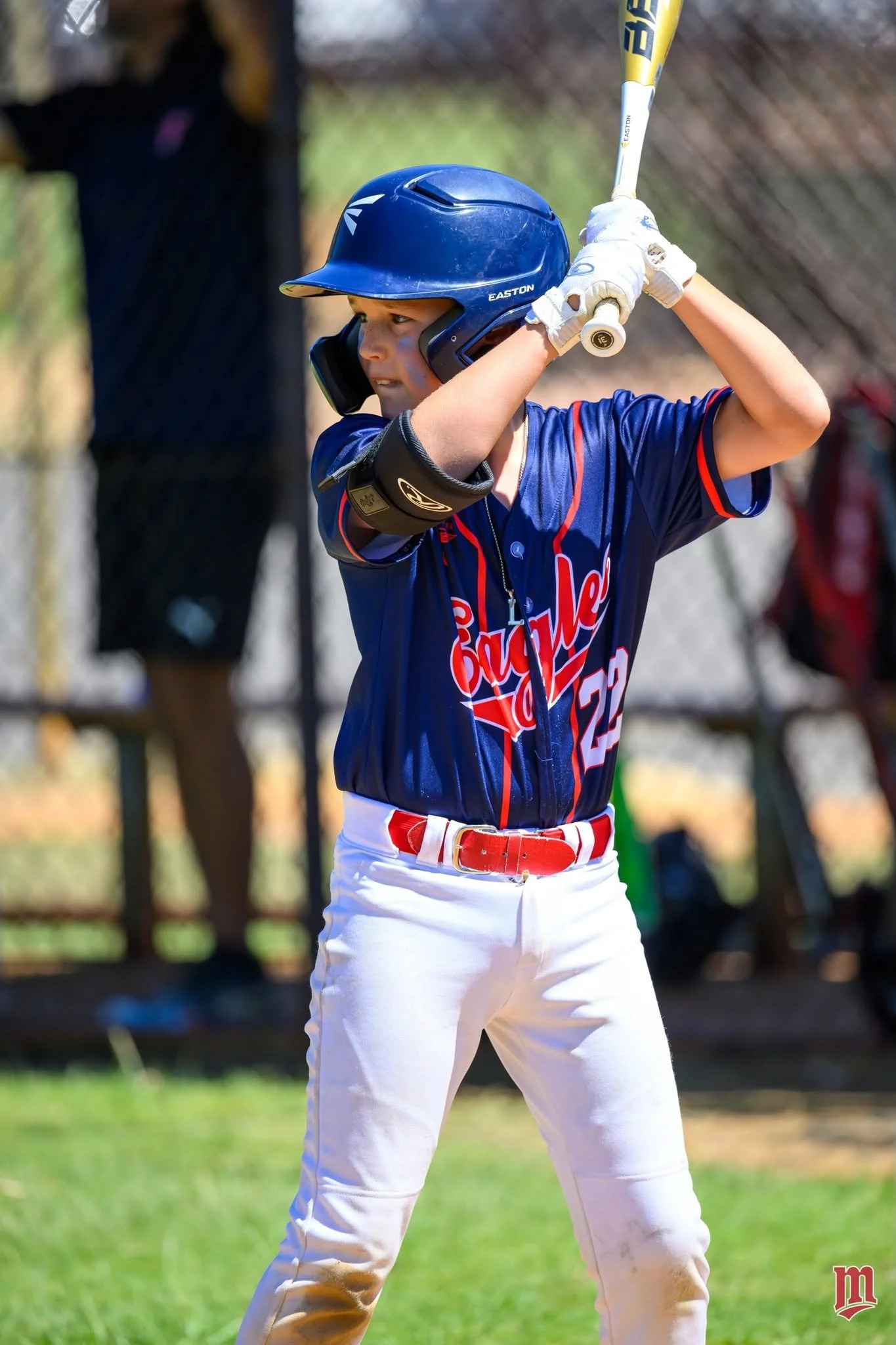 A young baseball player in a blue jersey and white pants swings a bat during a game, wearing a blue helmet and white gloves, with a green field and a chain-link fence in the background.