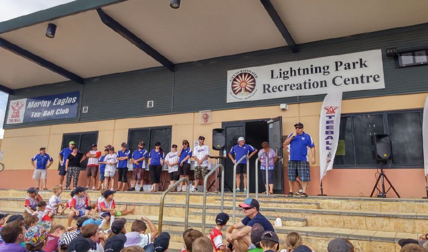 Children sitting on steps watching a group of people on an outdoor stage at Lightning Park Recreation Centre, with a sign for Morley Eagles Tee-Ball Club and a flag with 'Tee-Ball' on it.