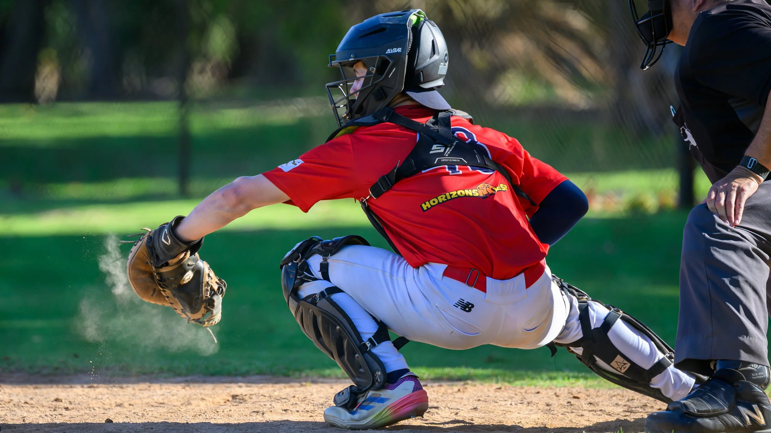 Baseball Catcher with red shirt and white pants squatting down to catch a ball in his glove. The catch creates a dust cloud in the glove.
