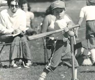 Young boy swinging a baseball bat at a ball on a tee during a game, with several people watching in the background.