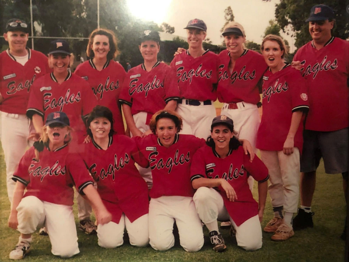 A group of young female softball players and coaches in red jerseys and white pants, posing on a field after a game.