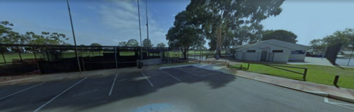 Empty parking lot near a sports field with trees and a building in the background.