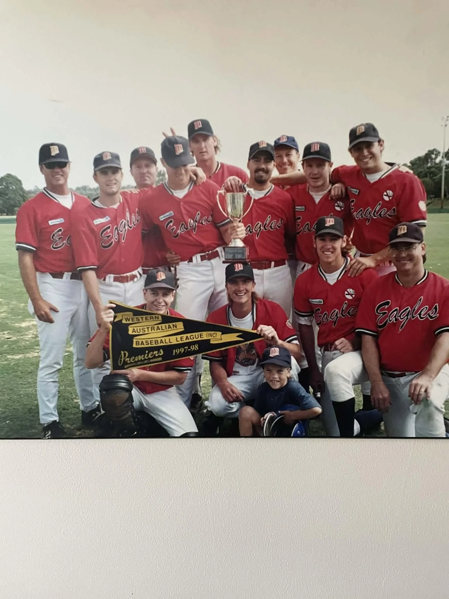 Group of baseball players wearing red jerseys and black caps, celebrating on a baseball field with a trophy.