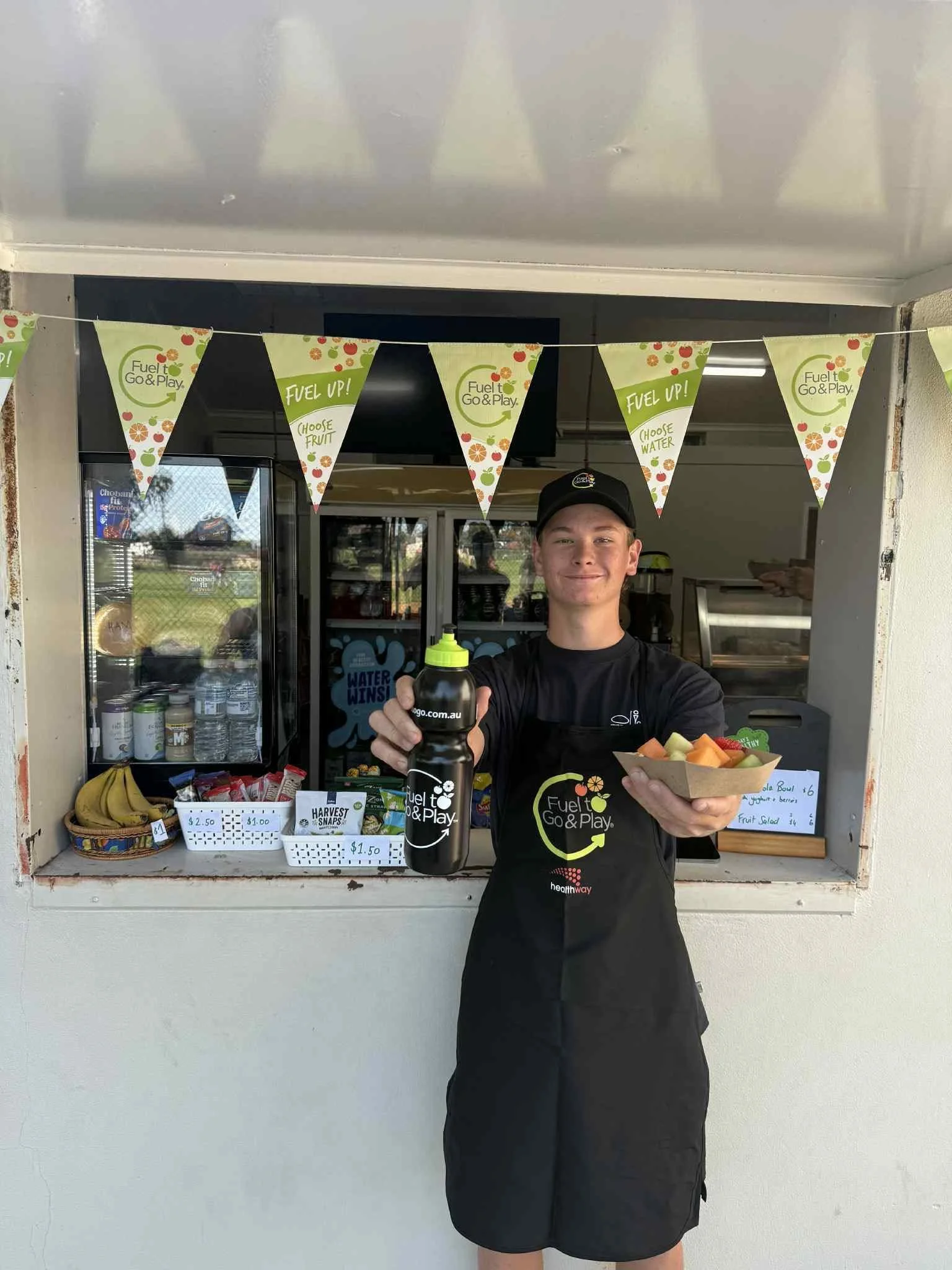 A young man wearing a black uniform standing in front of a small fruit stand, holding a water bottle and a bowl of sliced fruit, with a banner overhead that reads 'Fuel Up! Choose Fruit'.