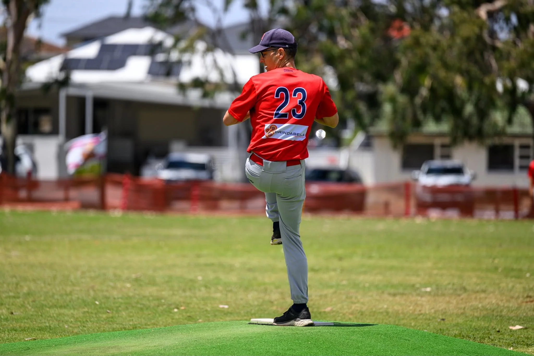 A person wearing a red sports jersey with the number 23, gray pants, wearing a cap and glasses, is standing on a green artificial surface with one foot on a round rubber base, preparing to pitch a game in a park or sports field.