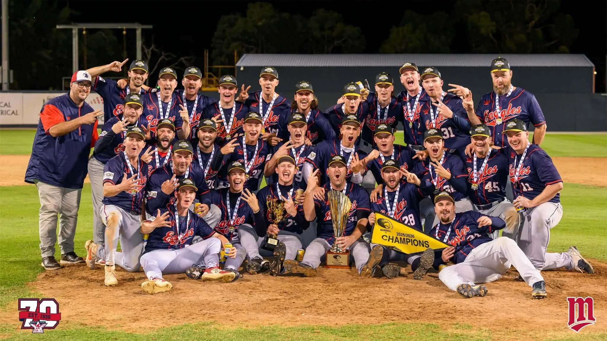 A group of baseball players and coaches celebrating on the field with trophies and medals, wearing navy and red uniforms, after winning a championship.