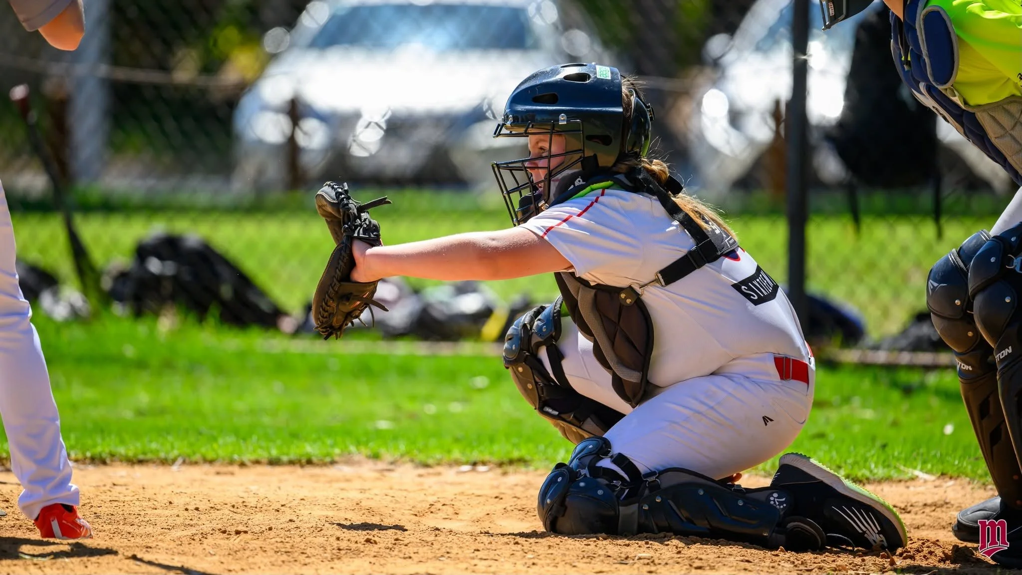 A girl playing baseball in a protective catcher's gear kneeling on the field, with the sunlight reflecting on her helmet and gear.