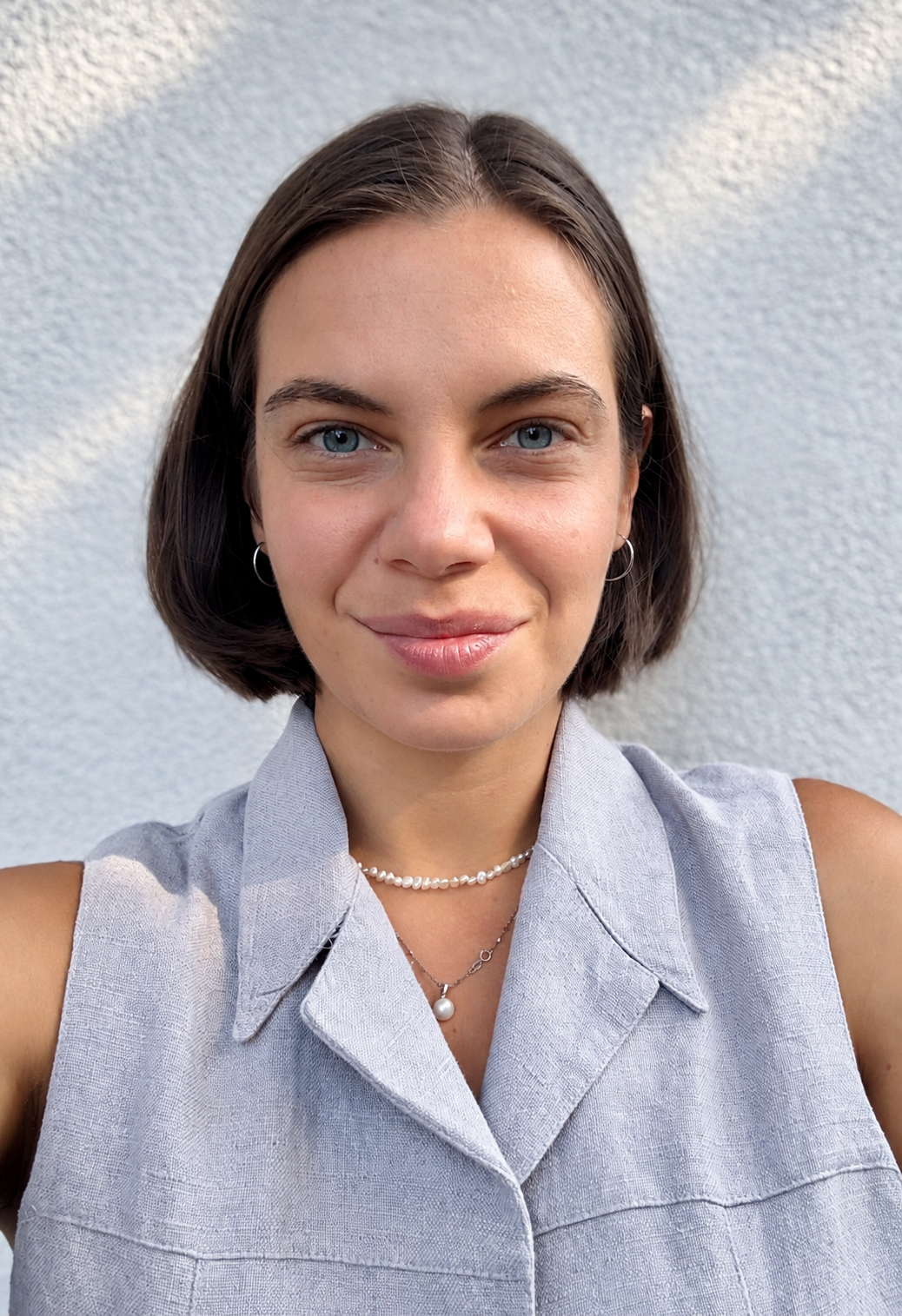 Portrait of a woman with short brown hair and blue eyes, smiling, wearing a sleeveless light gray collared shirt and layered pearl necklaces, standing against a light textured wall.