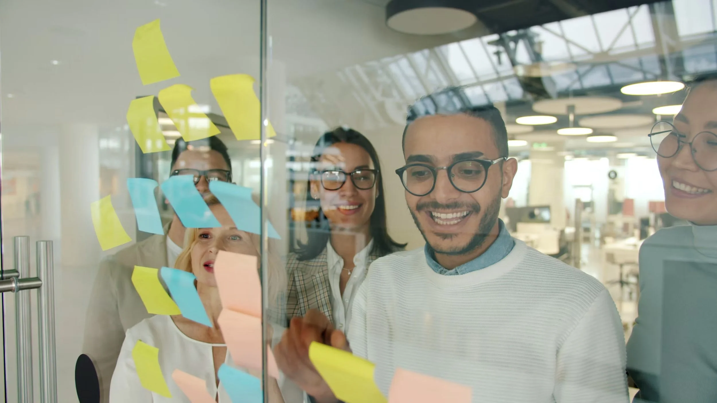 A diverse group of five young professionals gathered around a glass wall covered with colorful sticky notes, collaborating and smiling in a modern office environment with large windows and ceiling lights.
