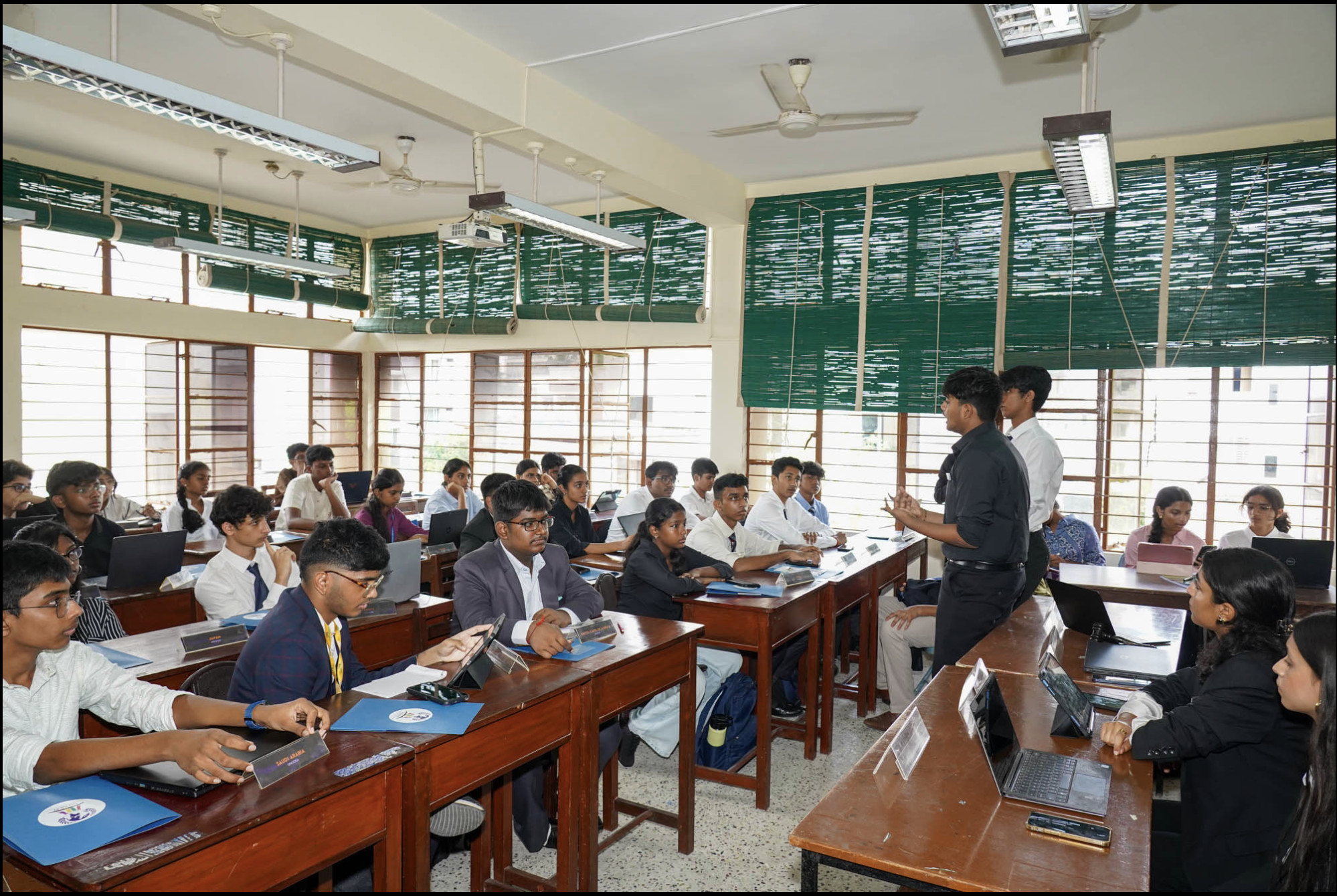 Students attending a classroom lecture, seated at desks with laptops and notebooks, listening to a teacher standing at the front.