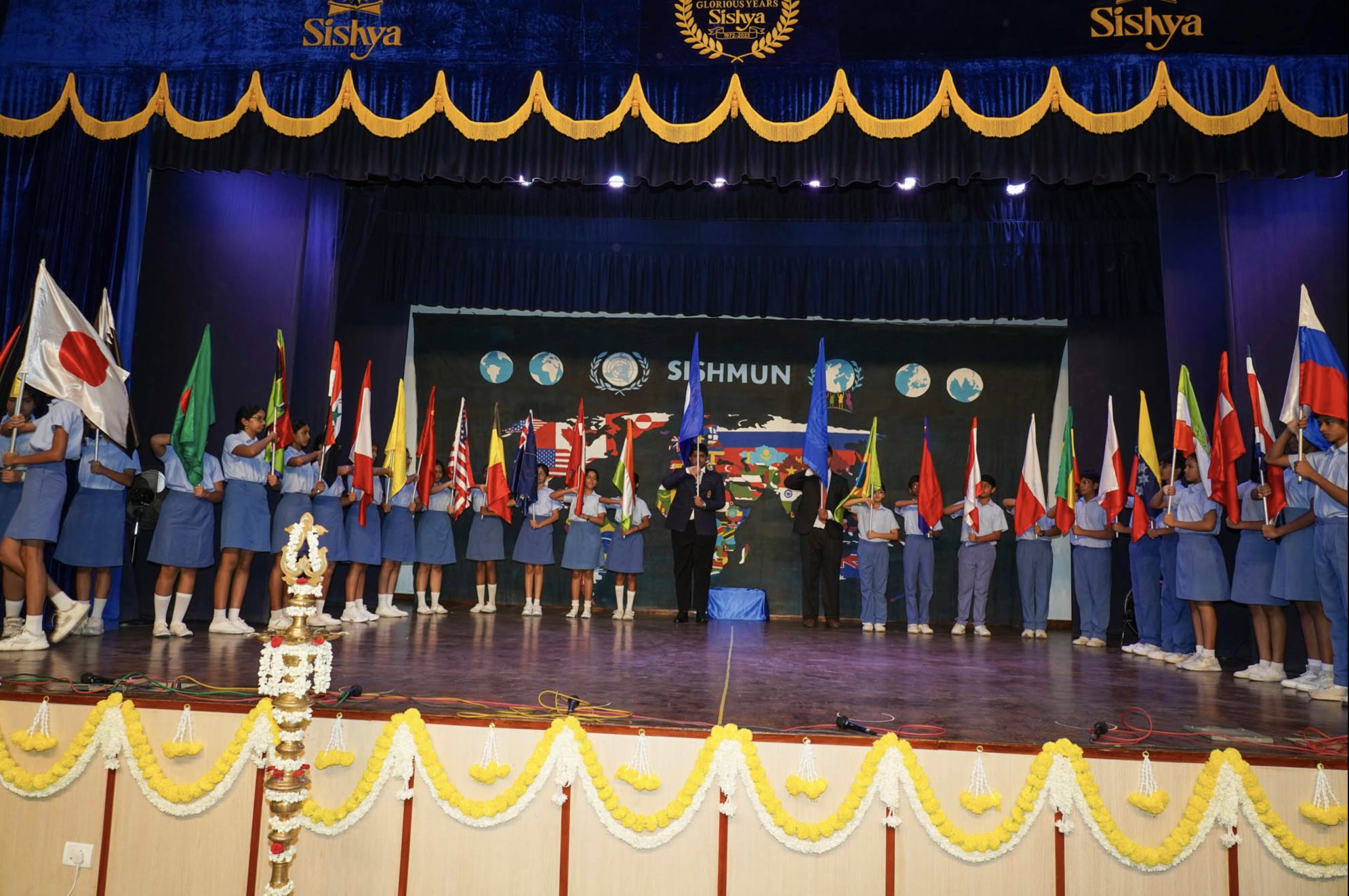 Students on stage participating in a cultural event holding international flags with floral decorations in front.