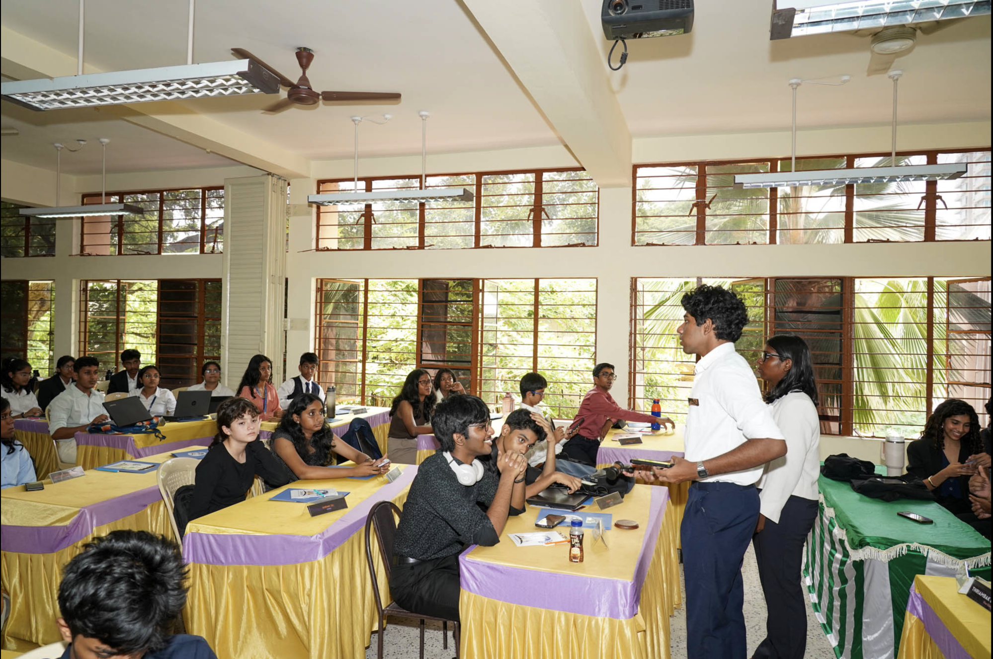 A classroom filled with students sitting at tables covered with yellow and purple tablecloths. Two educators, a man and a woman, are standing and engaging with the students. Large windows with open wooden shutters let in natural light, and lush greenery can be seen outside. The room is equipped with a ceiling fan, fluorescent lights, and a mounted projector.