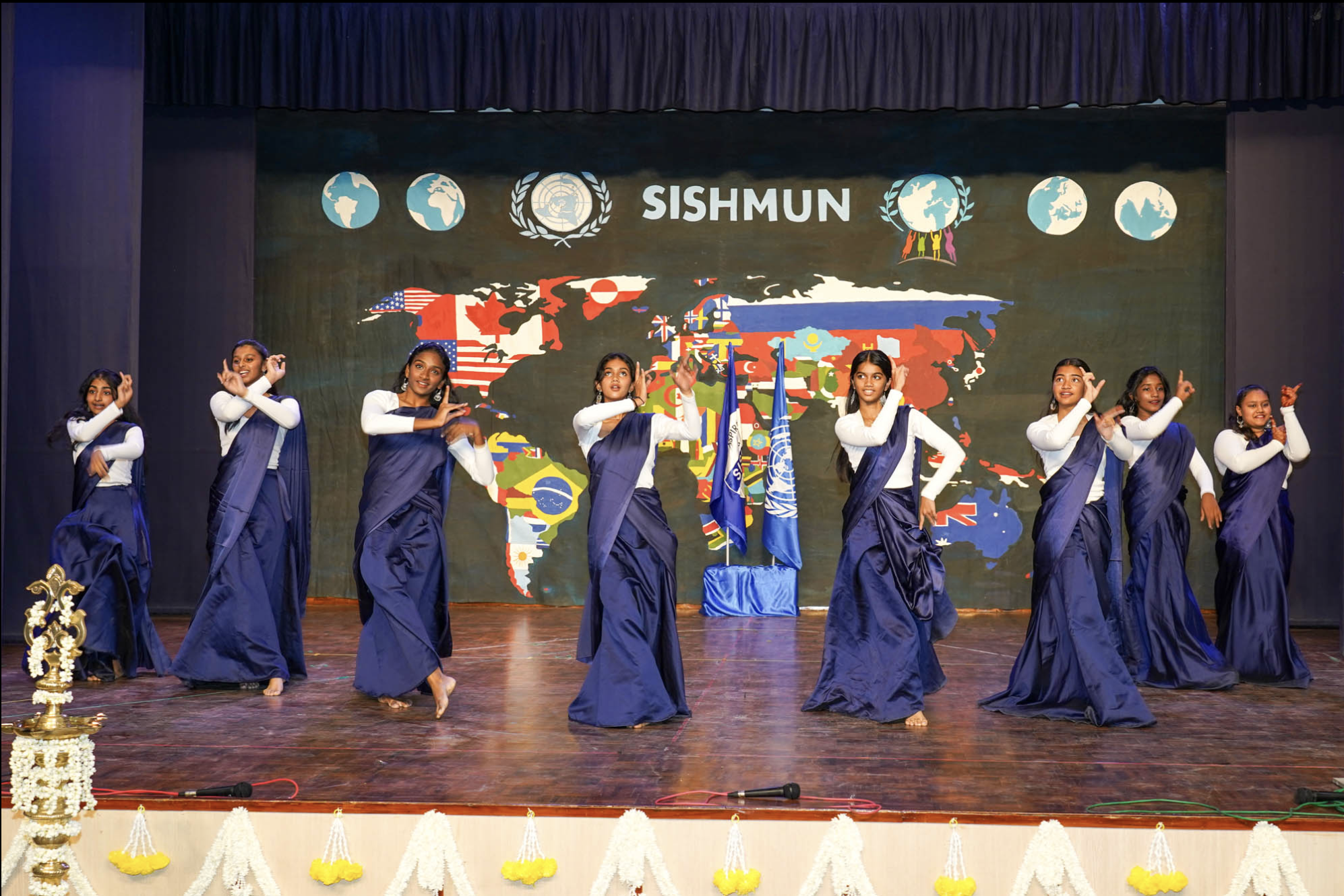 Group of young girls in traditional blue and white dresses performing a dance on stage, with a backdrop featuring a map of the world and symbols representing international unity. There are flags and a decorated platform on stage.