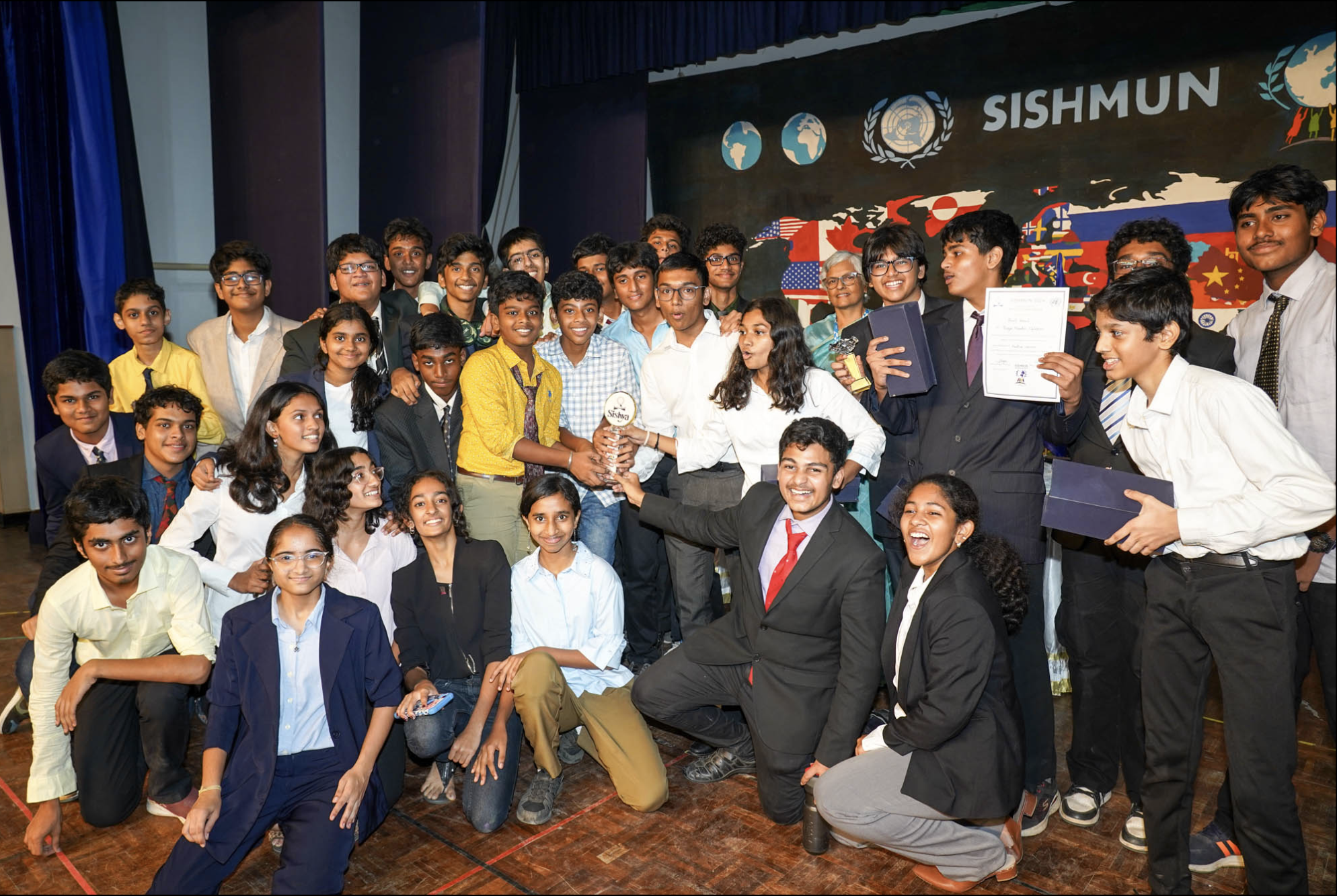 Group of young students and teachers on a stage celebrating a competition or award ceremony, with a World Map backdrop featuring country flags and the SISHMUN logo.