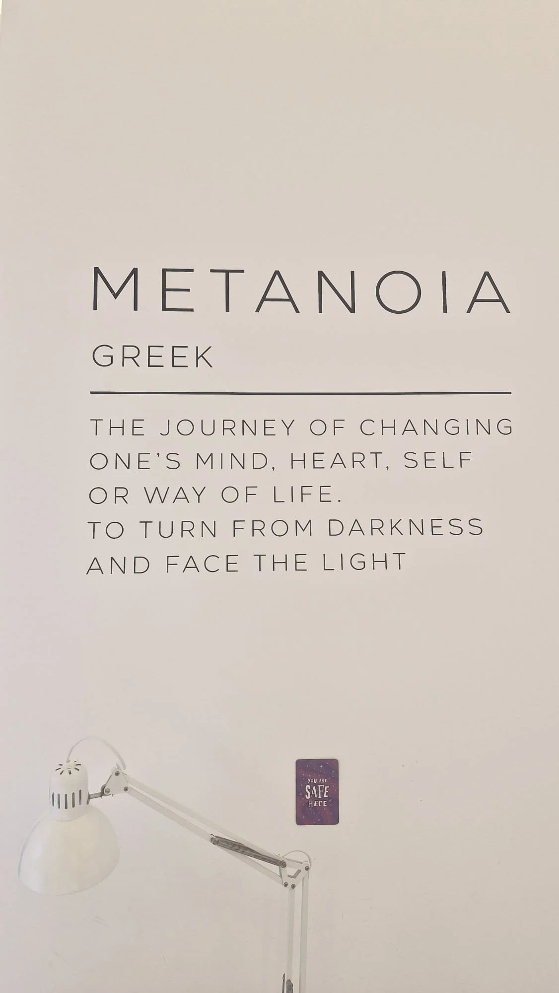 A wall poster titled 'Metanoia Greek' with a quote about changing one's mind, heart, self, or way of life to face the light, a white adjustable lamp, and a small purple sign that says 'You are safe here'.