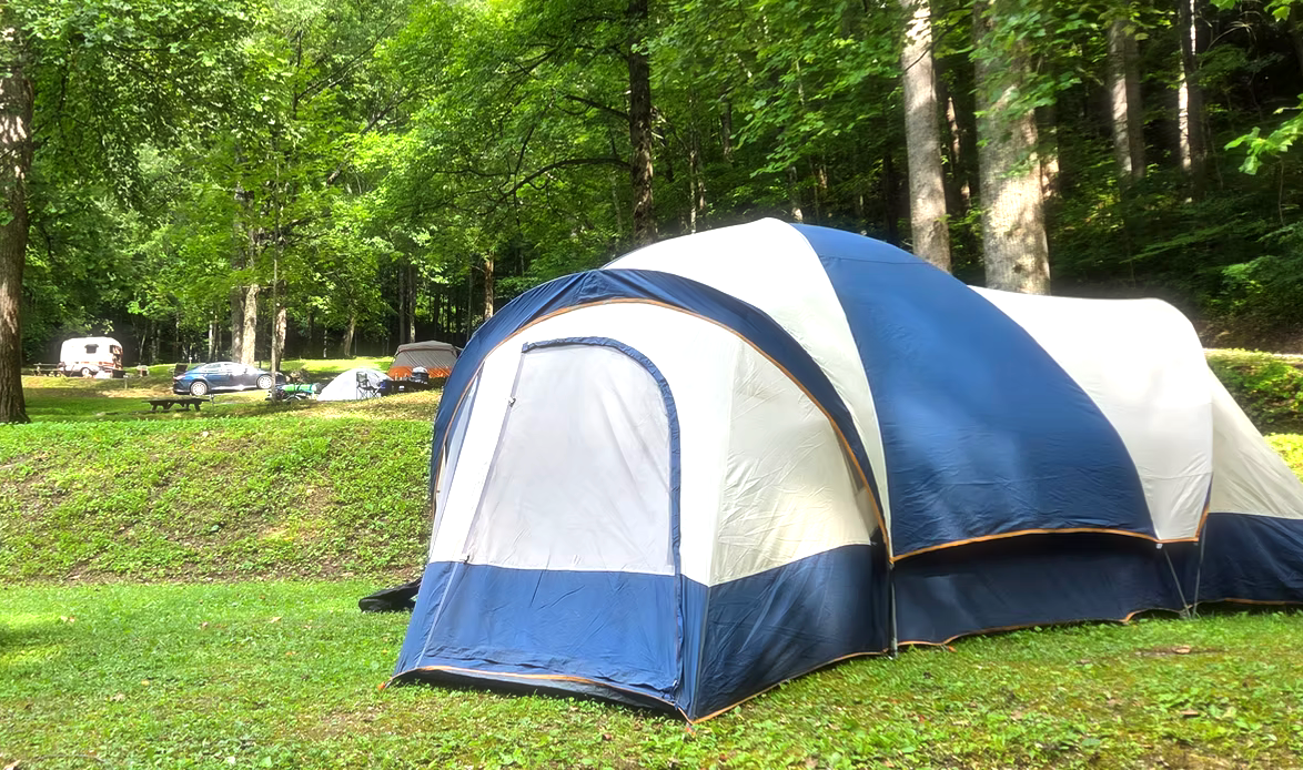Large blue and white family tent at a wooded campground with cars, picnic tables, and other tents in the background.