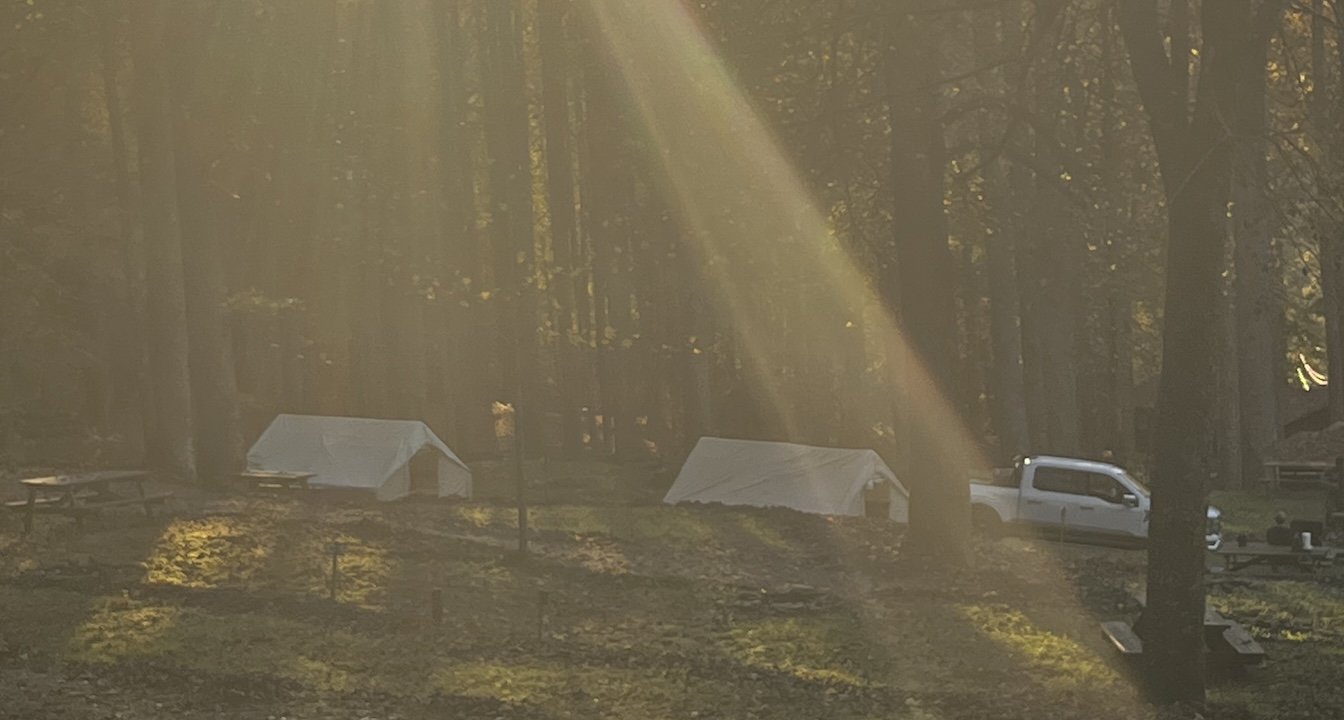 Sunbeams shine through a forest over two white camping tents, picnic tables, and a parked pickup truck.