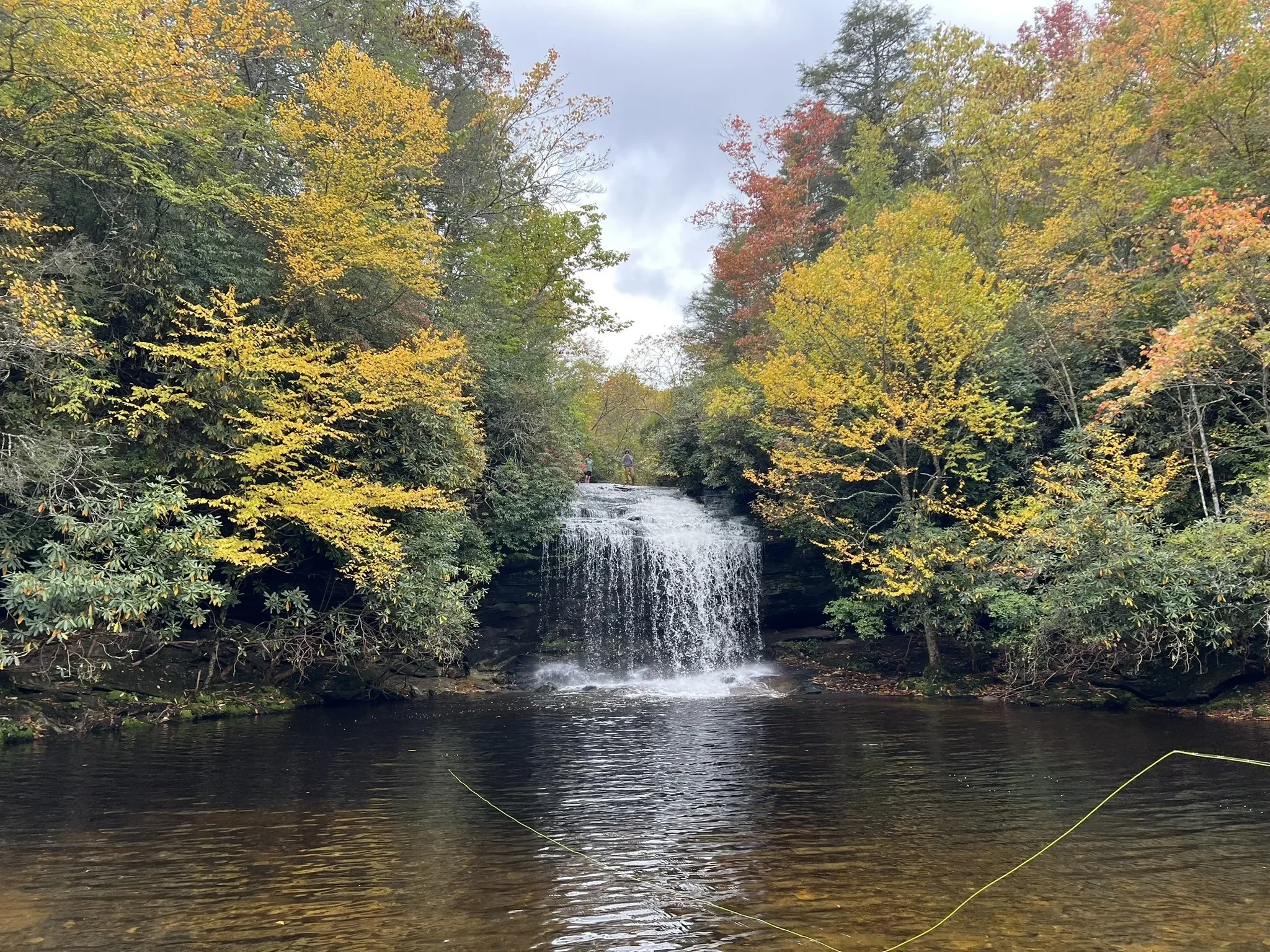 Waterfall flowing into a calm pool surrounded by colorful autumn forest, with two people standing above the falls.