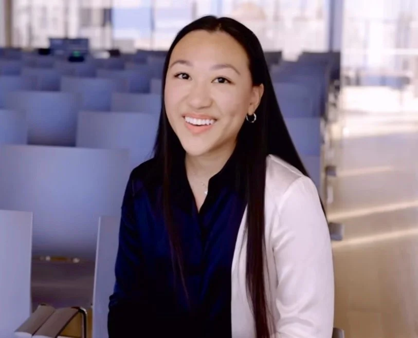 A young woman with long dark hair, earrings, and a bright smile, sitting in an empty conference room with rows of blue chairs and large windows.