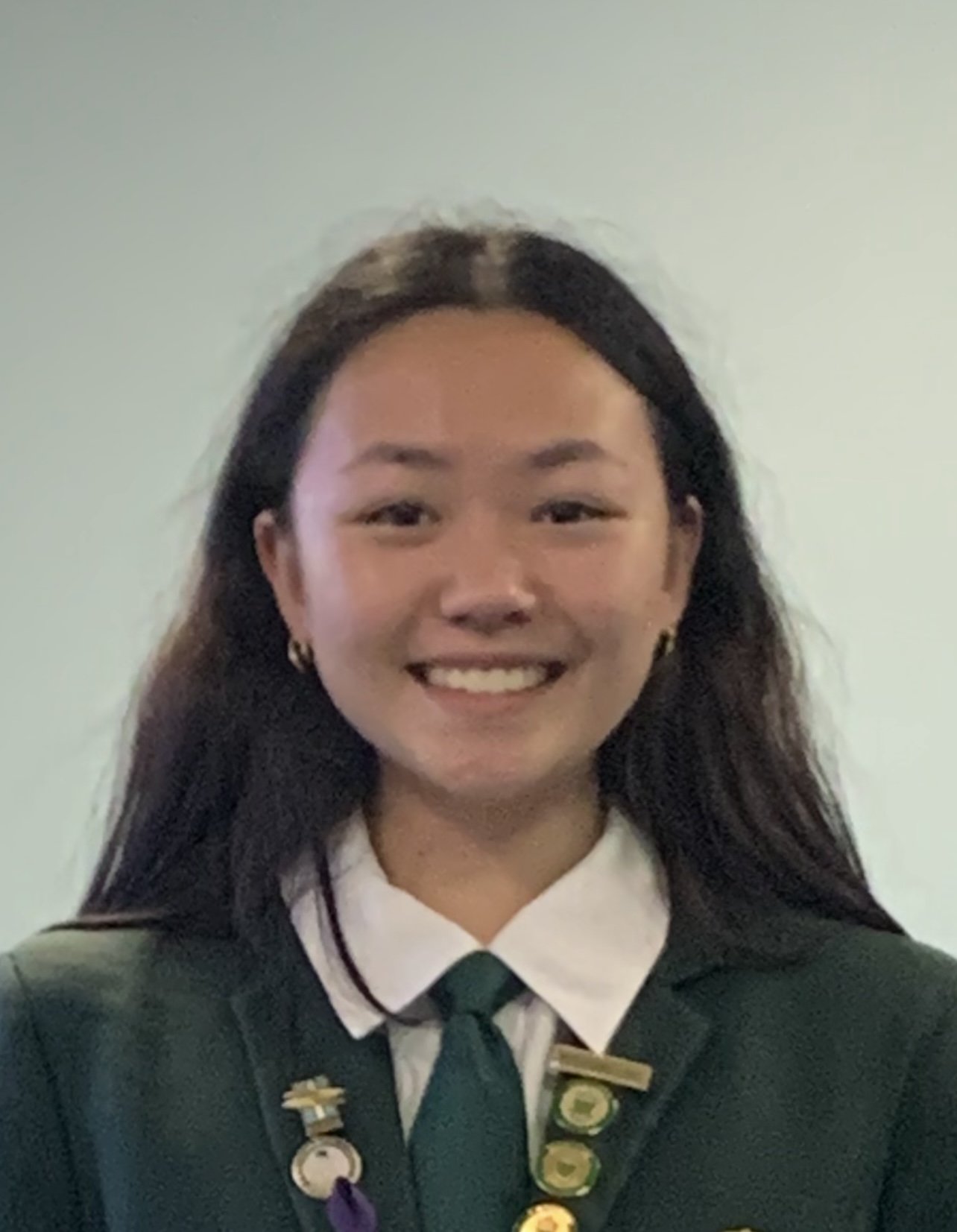 Young woman with long dark hair in a green school uniform with a white shirt, green tie, and several badges, smiling against a light-colored wall.