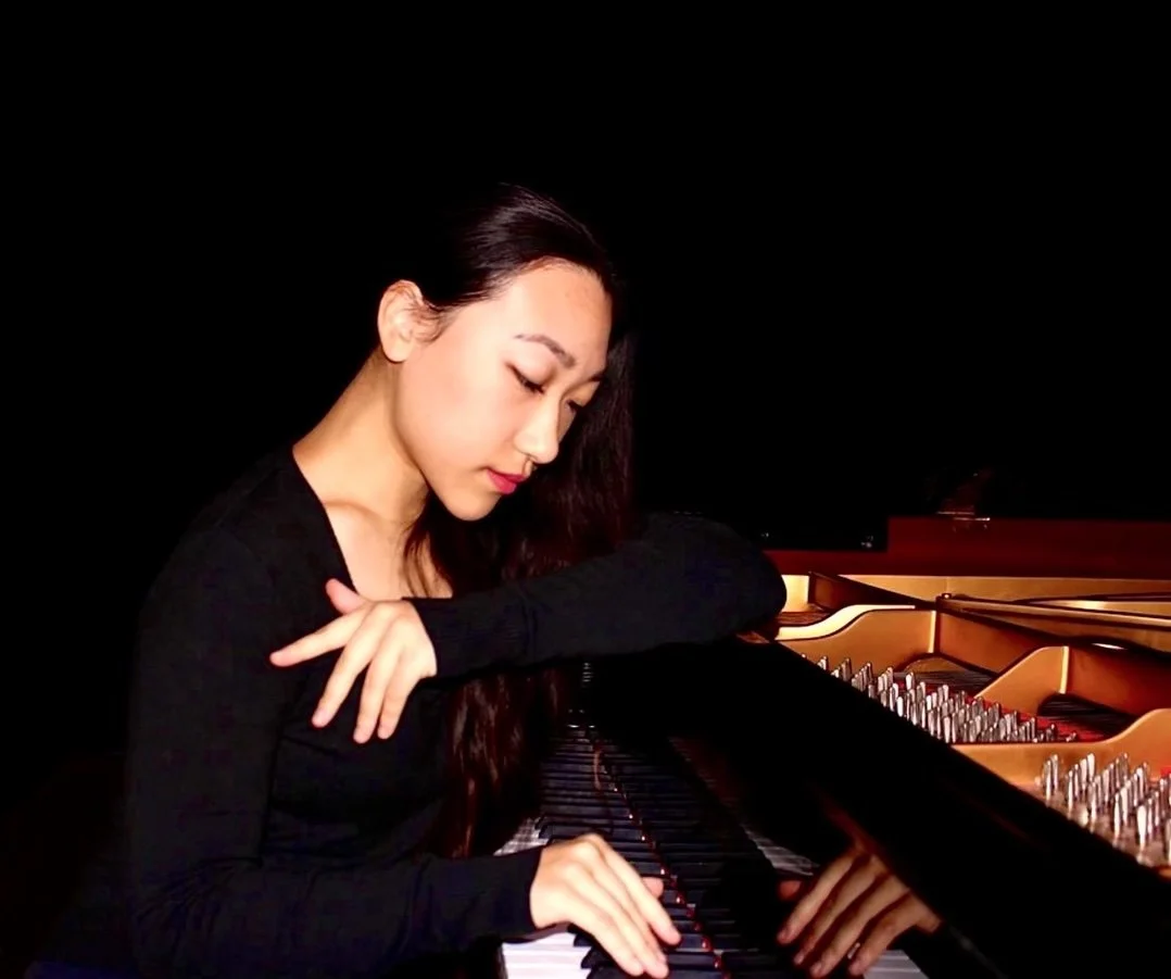 Young woman with long dark hair playing a grand piano in a dark setting.