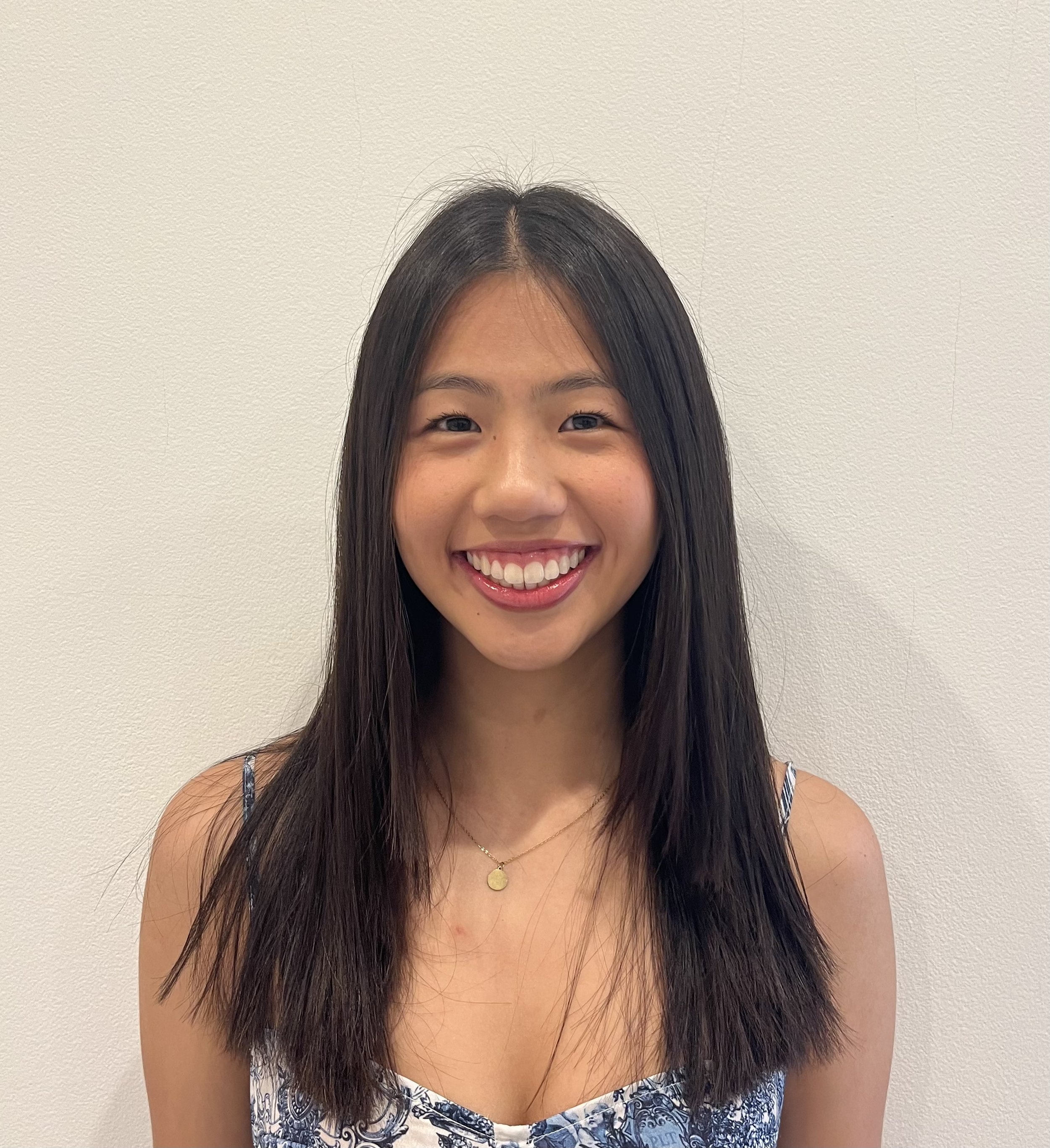 A young woman with long dark hair smiling in front of a plain white wall, wearing a patterned dress and a gold necklace.