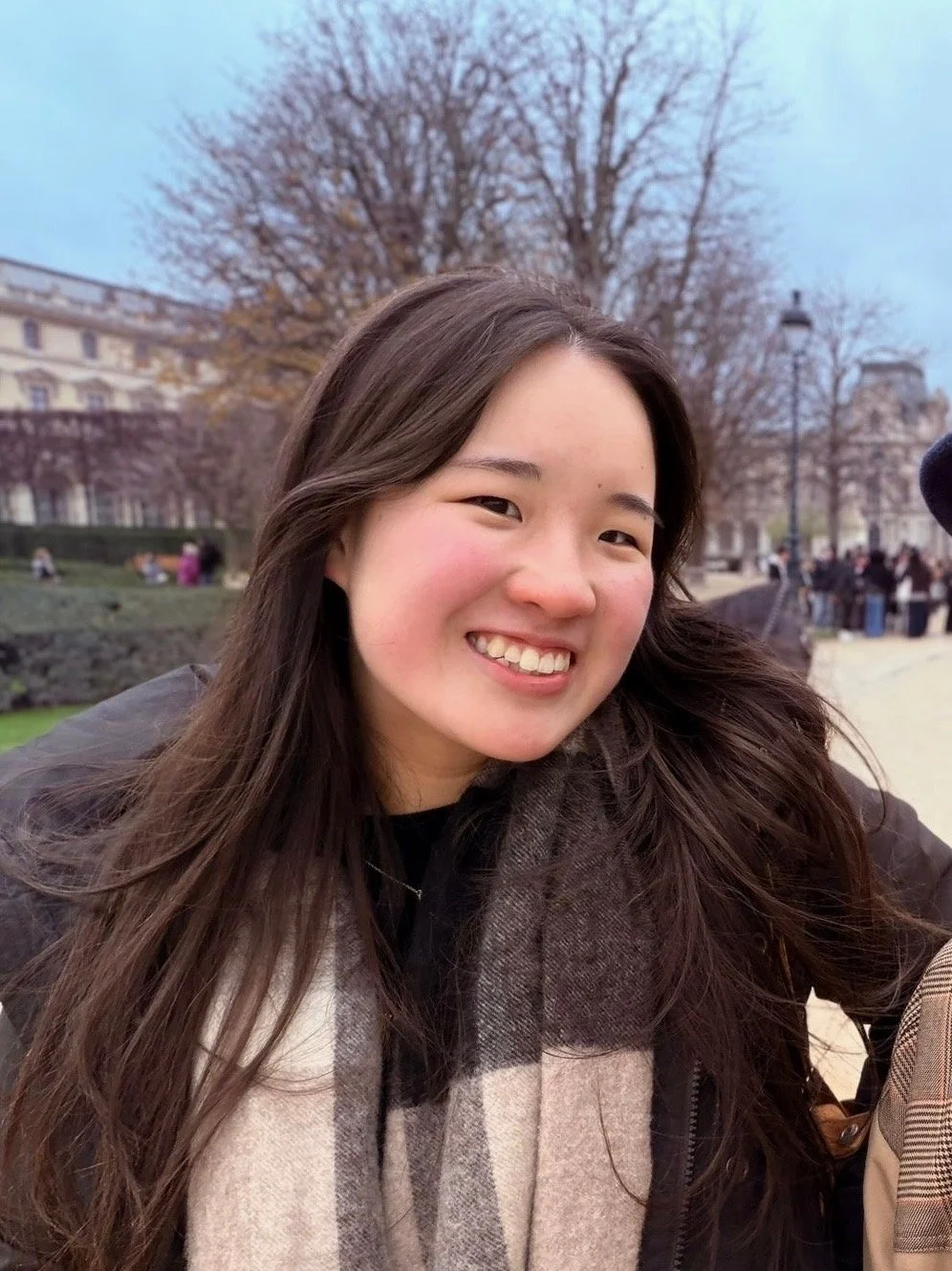 A young woman with long brown hair smiling outdoors in a park with leafless trees and historic buildings in the background.