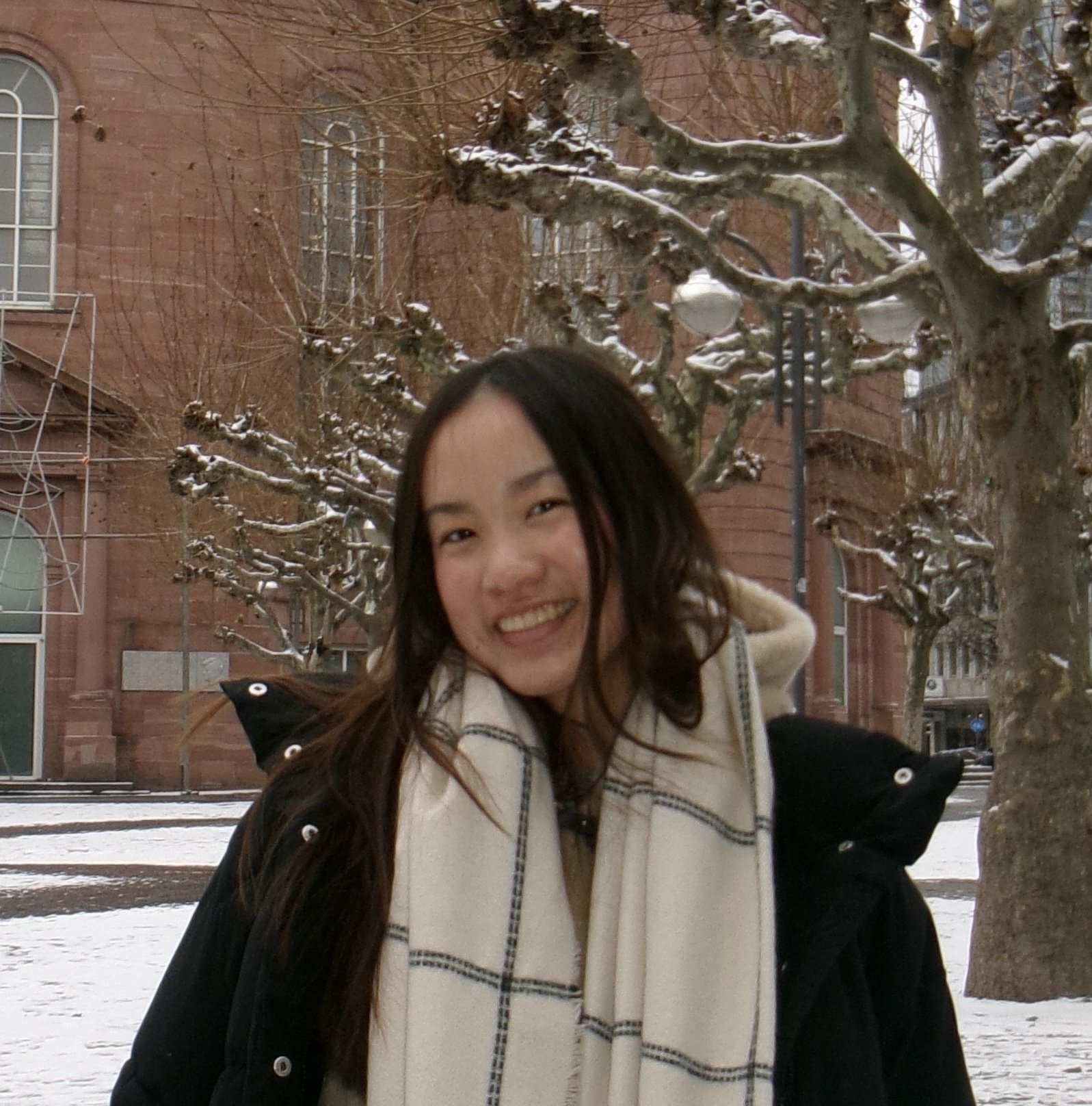 A smiling woman with long dark hair posing outdoors in a snowy urban setting, with leafless trees and brick buildings in the background.
