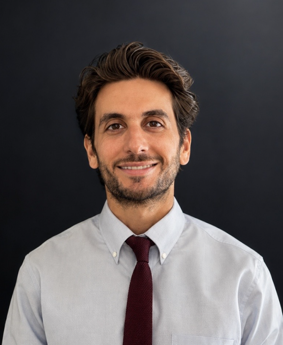 A young man with dark brown, wavy hair and a beard, smiling, wearing a light gray button-up shirt and a dark red tie, standing against a dark background.
