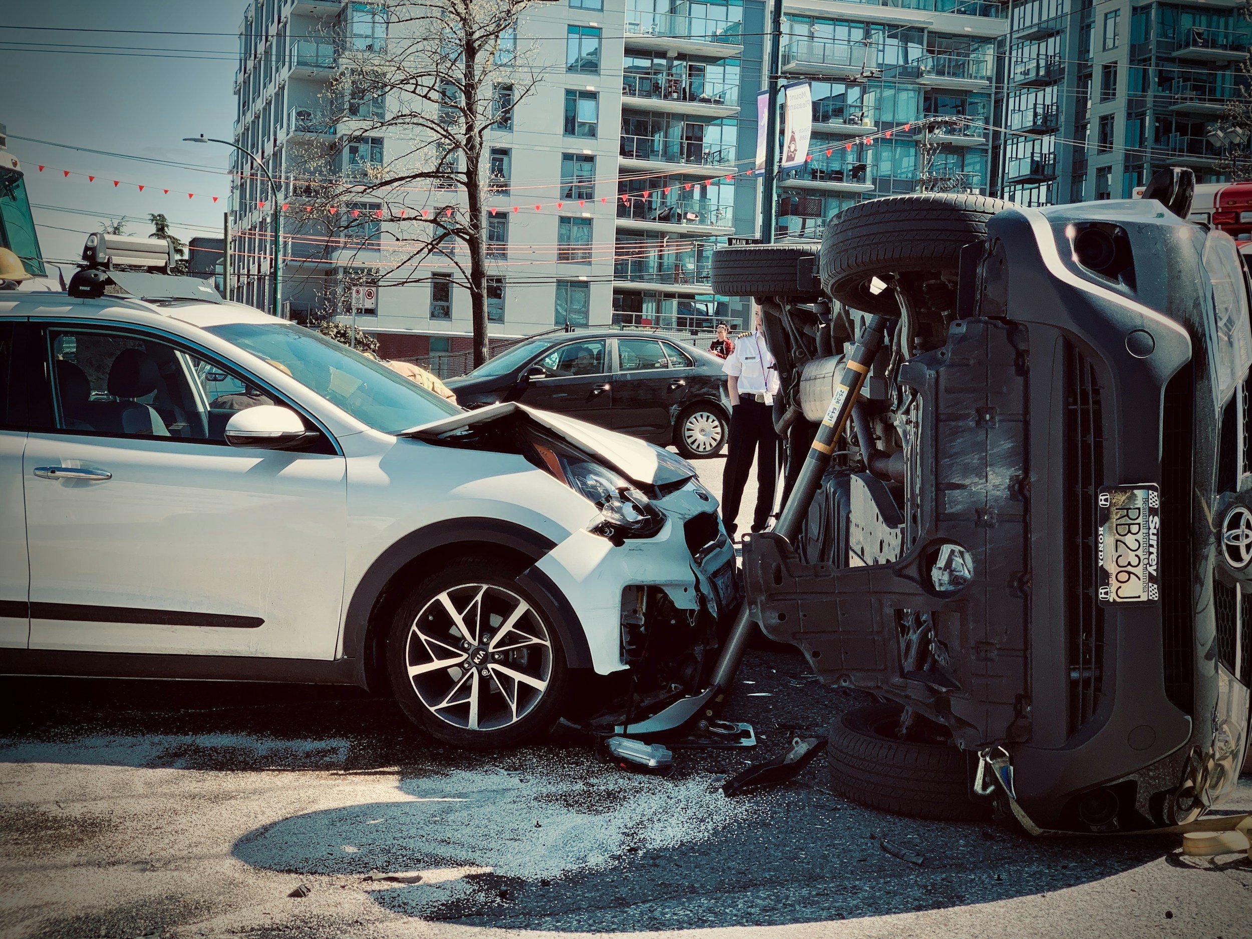 A car accident scene showing a white car with front-end damage and a black SUV on its side. The accident takes place in an urban area with high-rise apartment buildings in the background.