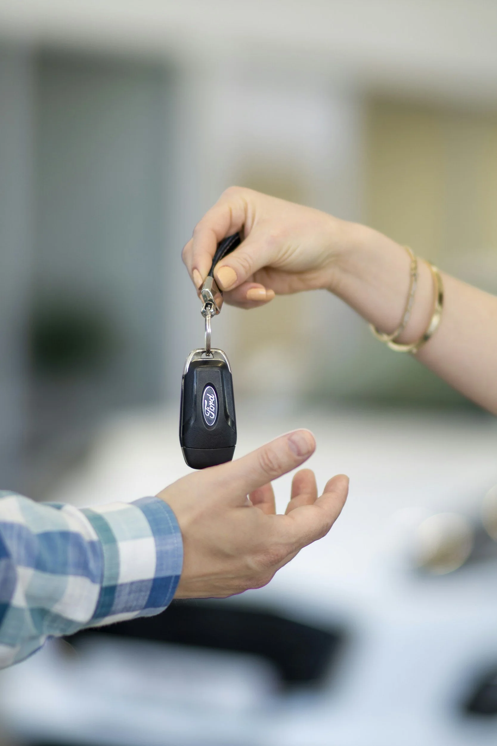 A person hands a car key to another person, who is wearing a blue and white checkered shirt.