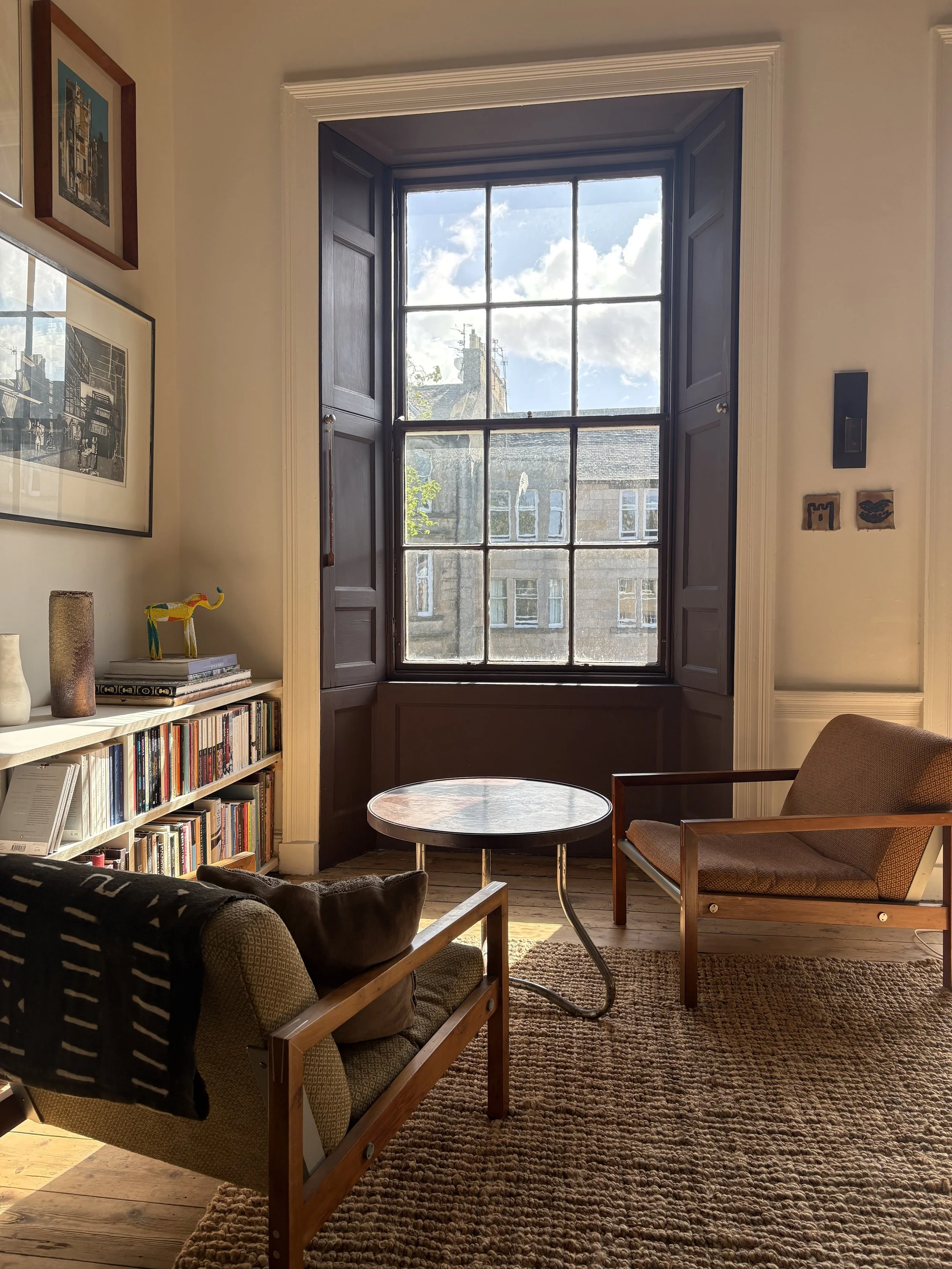 Interior of a cozy living room with a large window, wooden floor, and a mix of modern and vintage furniture including a lounge chair with a wooden frame and patterned upholstery, a small round table, and a bookshelf filled with books. Decor includes vases, framed artwork, and small sculptures. Natural light streams through the window revealing neighboring buildings and a blue sky with clouds.