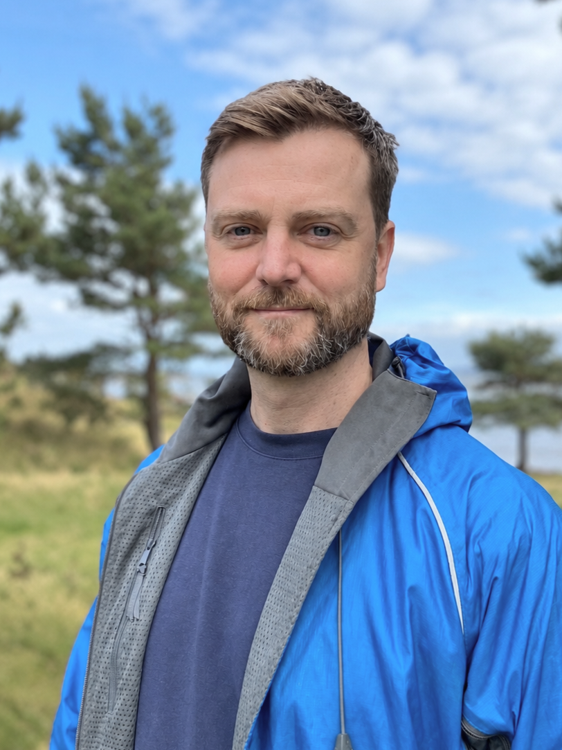 Portrait of a bearded man wearing a bright blue outdoor jacket near a lake and pine trees