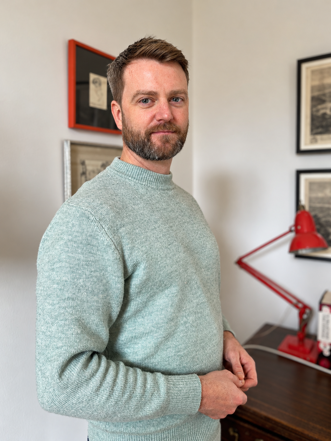A man with short brown hair and a beard standing in a room with framed pictures on the wall, wearing a light green sweater, next to a red desk lamp.