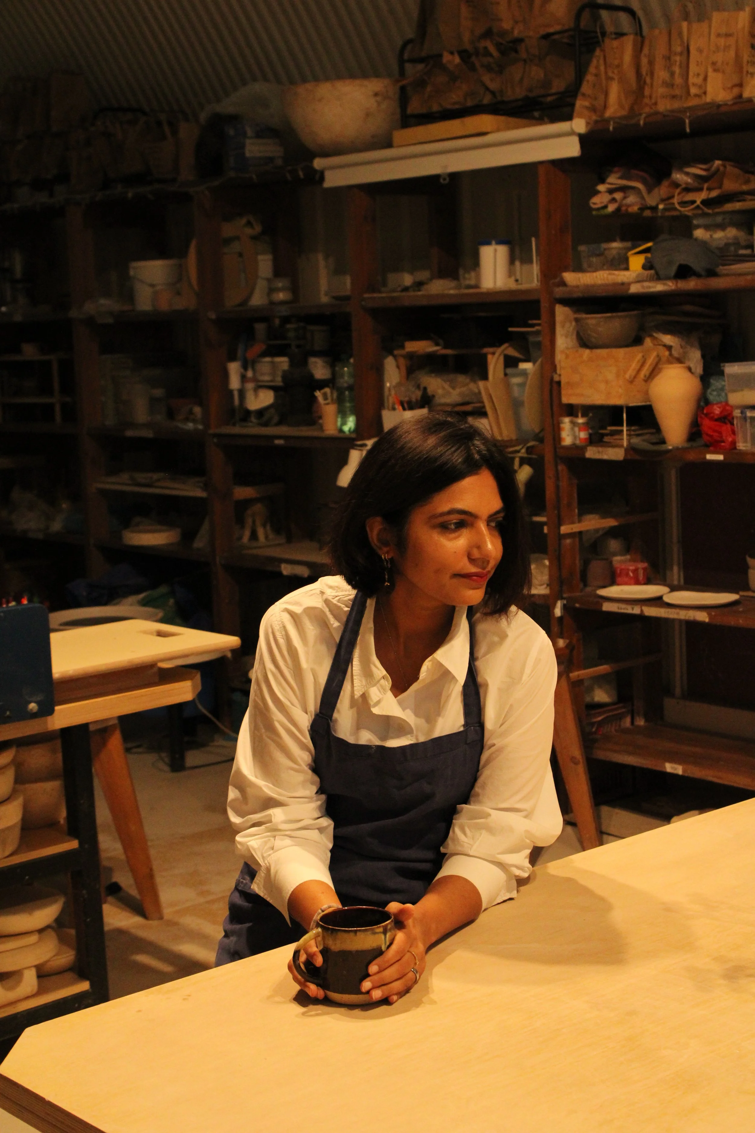 A woman with dark hair and a beige shirt wearing a black apron, sitting at a light-colored wooden table holding a black mug, in a workshop or studio with wooden shelves filled with pottery, tools, and supplies in the background.
