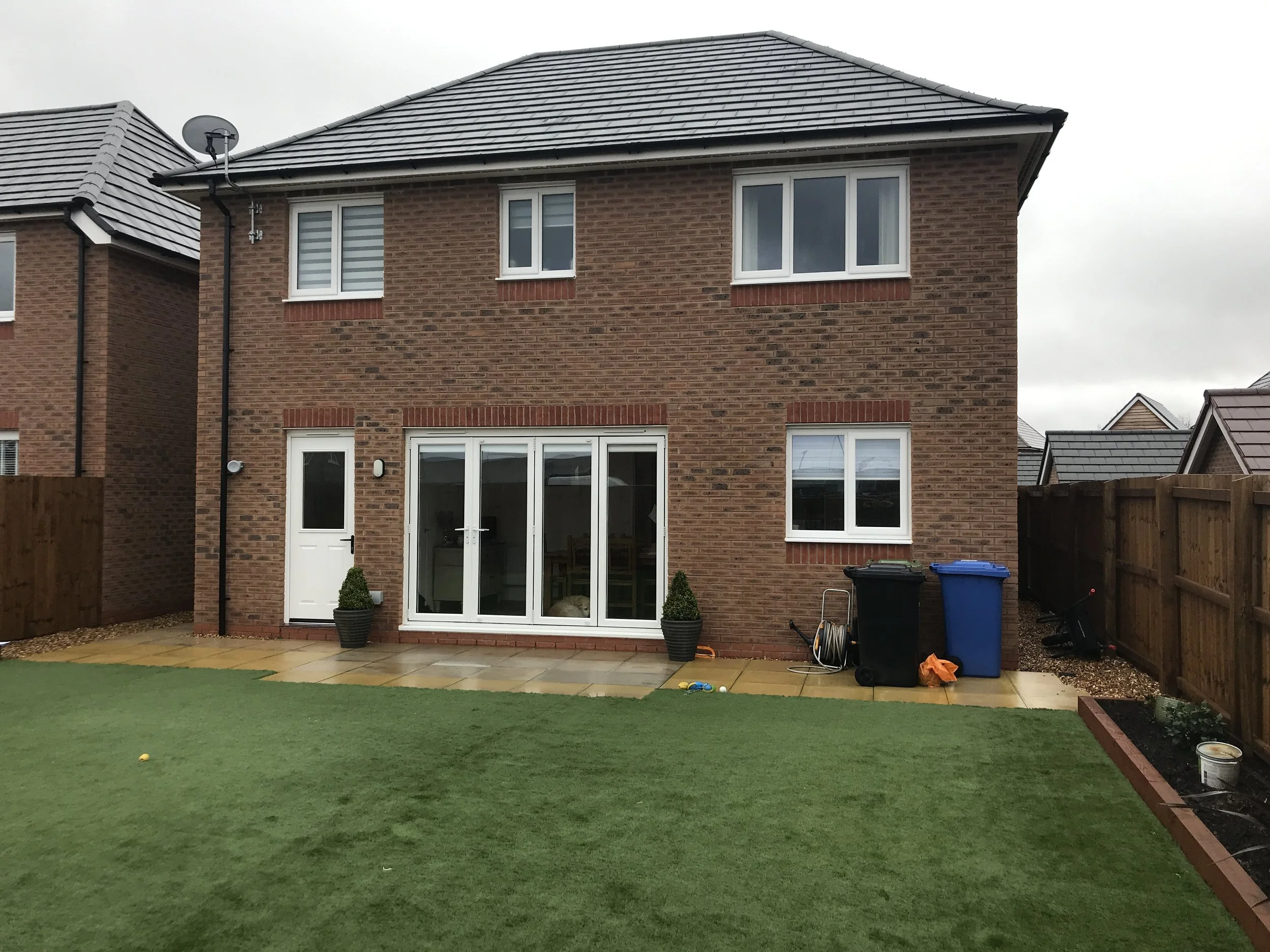 Backyard view of a two-story brick house with a sliding glass door, windows, and a patio area. The yard has artificial green grass, a small garden bed, and some outdoor trash bins. Overcast sky.