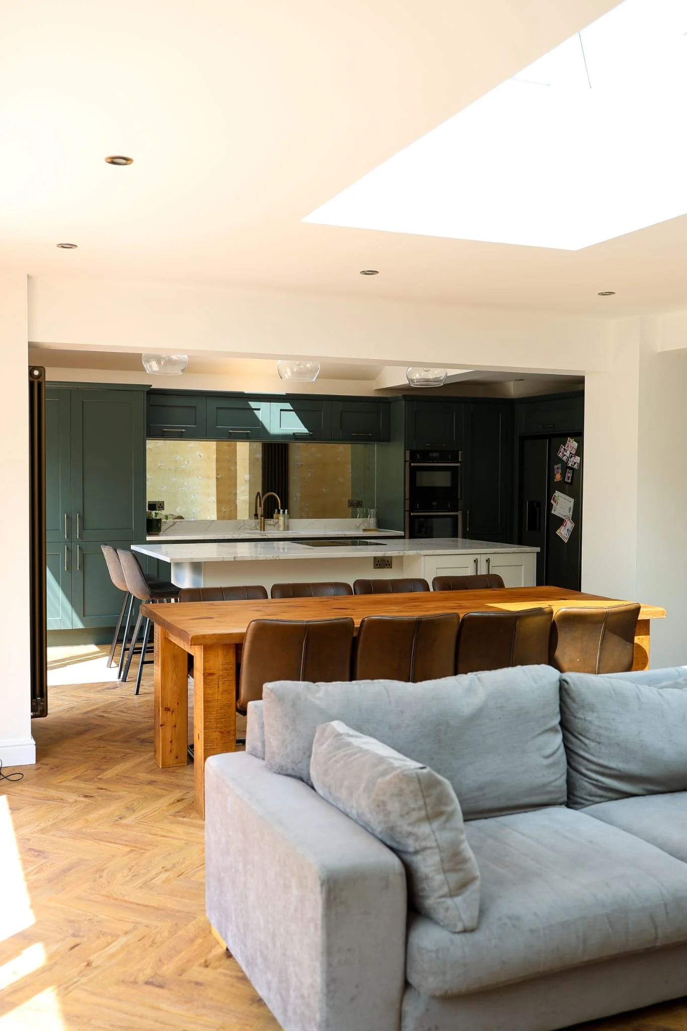 Modern kitchen with dark green cabinets, white island, gold fixtures, and a ceiling skylight, seen from a living room with a light-colored sofa and a wooden dining table.