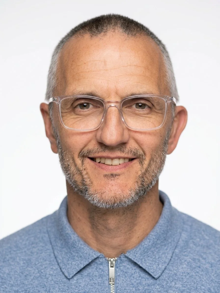 Close-up portrait of a middle-aged man with glasses, gray hair, and a beard, smiling and wearing a blue zipped jacket.