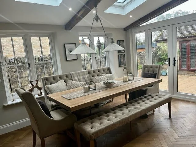 Dining room with a wooden table, upholstered chairs, and a matching bench, lit by pendant lights, with large windows and sliding glass doors showing an outdoor patio.