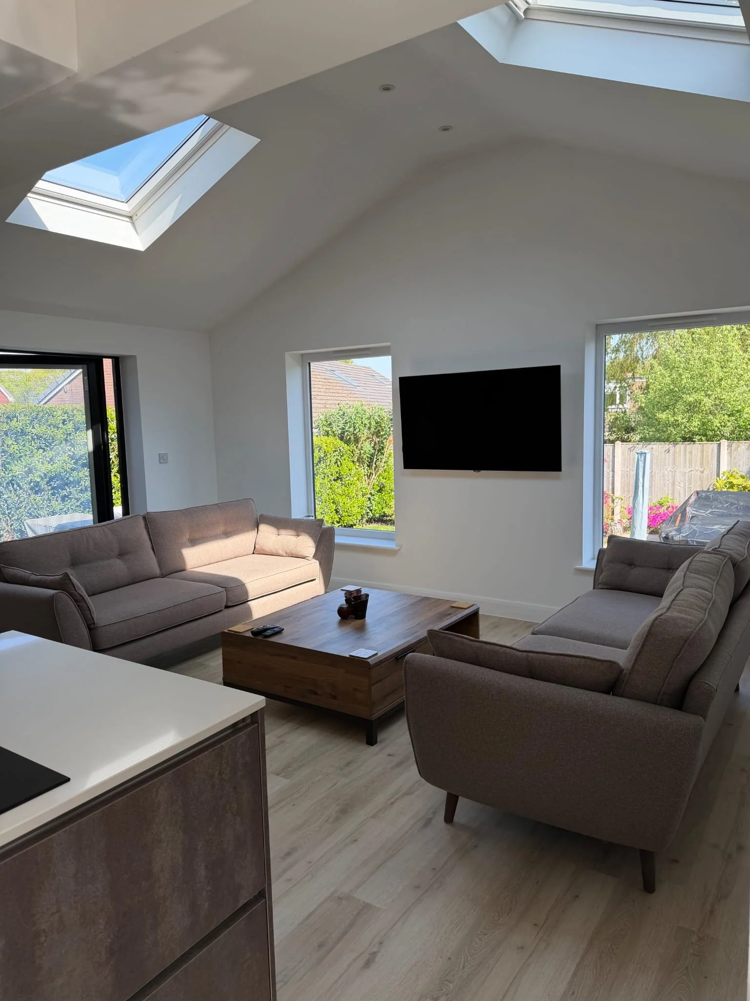 Living room with two beige couches, a wooden coffee table, flat-screen TV on the wall, and windows showing a garden outside with trees and fencing. Skylights in the ceiling let in natural light.