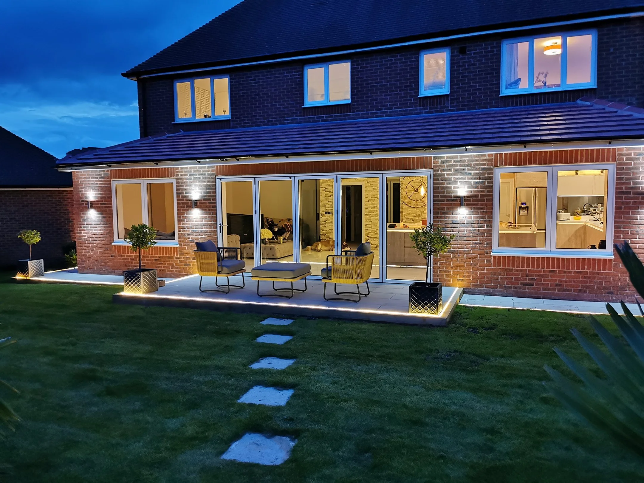A two-story house with brick exterior and large windows, illuminated from the inside, with a backyard patio area featuring outdoor furniture and potted plants, during dusk or early evening.
