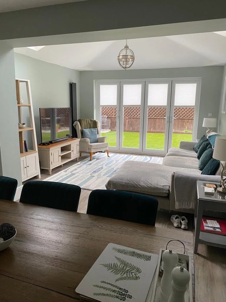 Living room with glass double doors leading to a backyard, a beige armchair, a large flat-screen TV on a stand, a patterned area rug, a white sofa with teal pillows, and a dining table partially visible in the foreground.