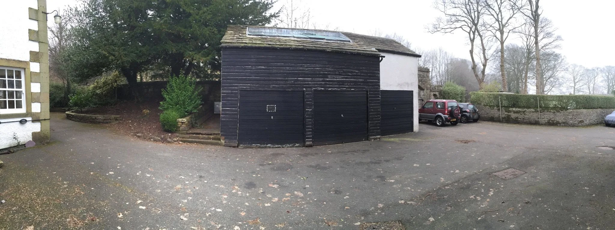 A small building with a black wooden exterior, two garage doors, and a skylight on the roof. To the right, there are parked cars along a stone wall, with a hedge and leafless trees in the background. To the left, part of a white building and a garden