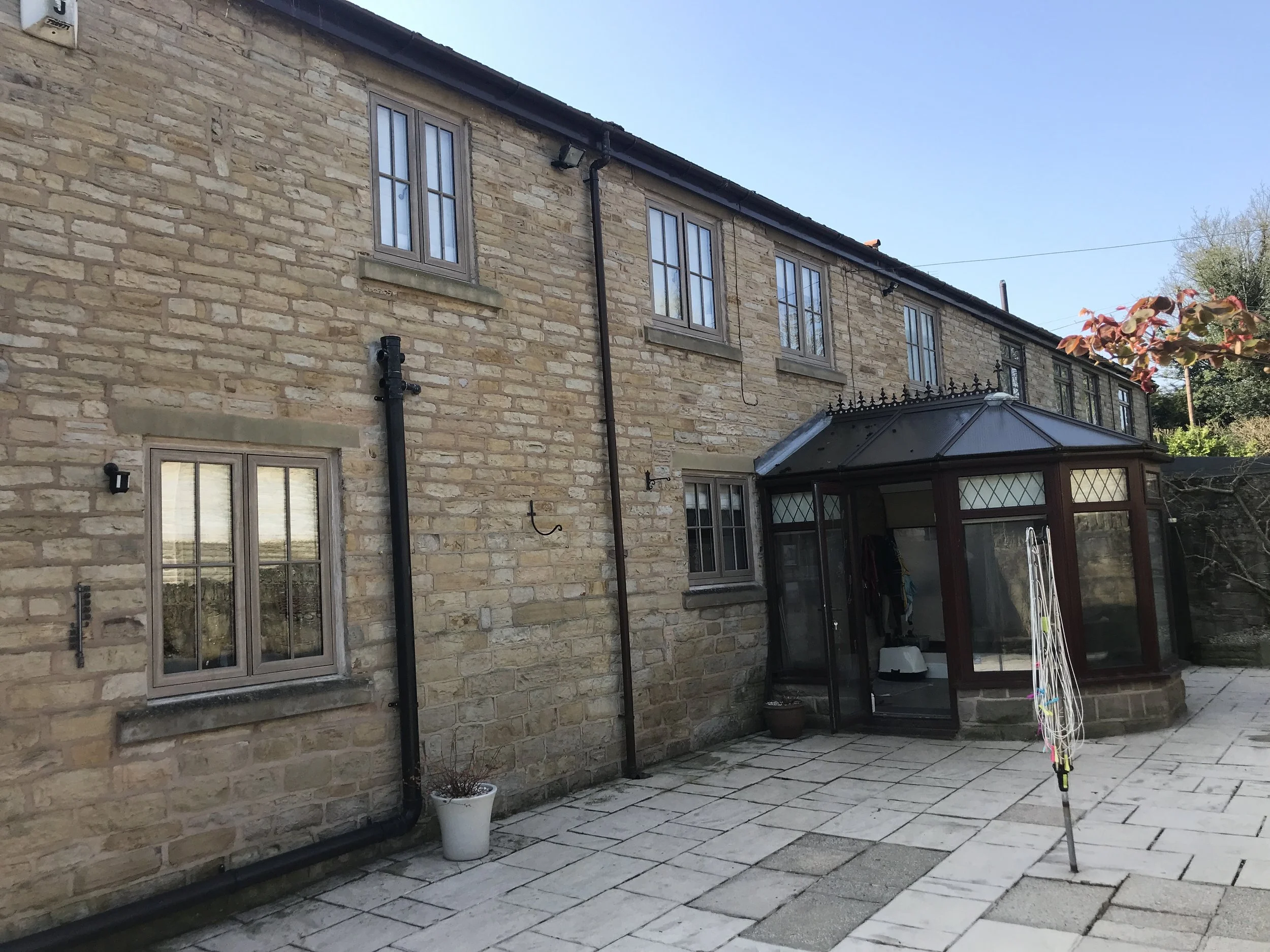 Backyard patio area with stone paving, a brick house wall with several windows, a potted plant, and a glass conservatory. A clothes drying pole and some outdoor equipment are also visible.