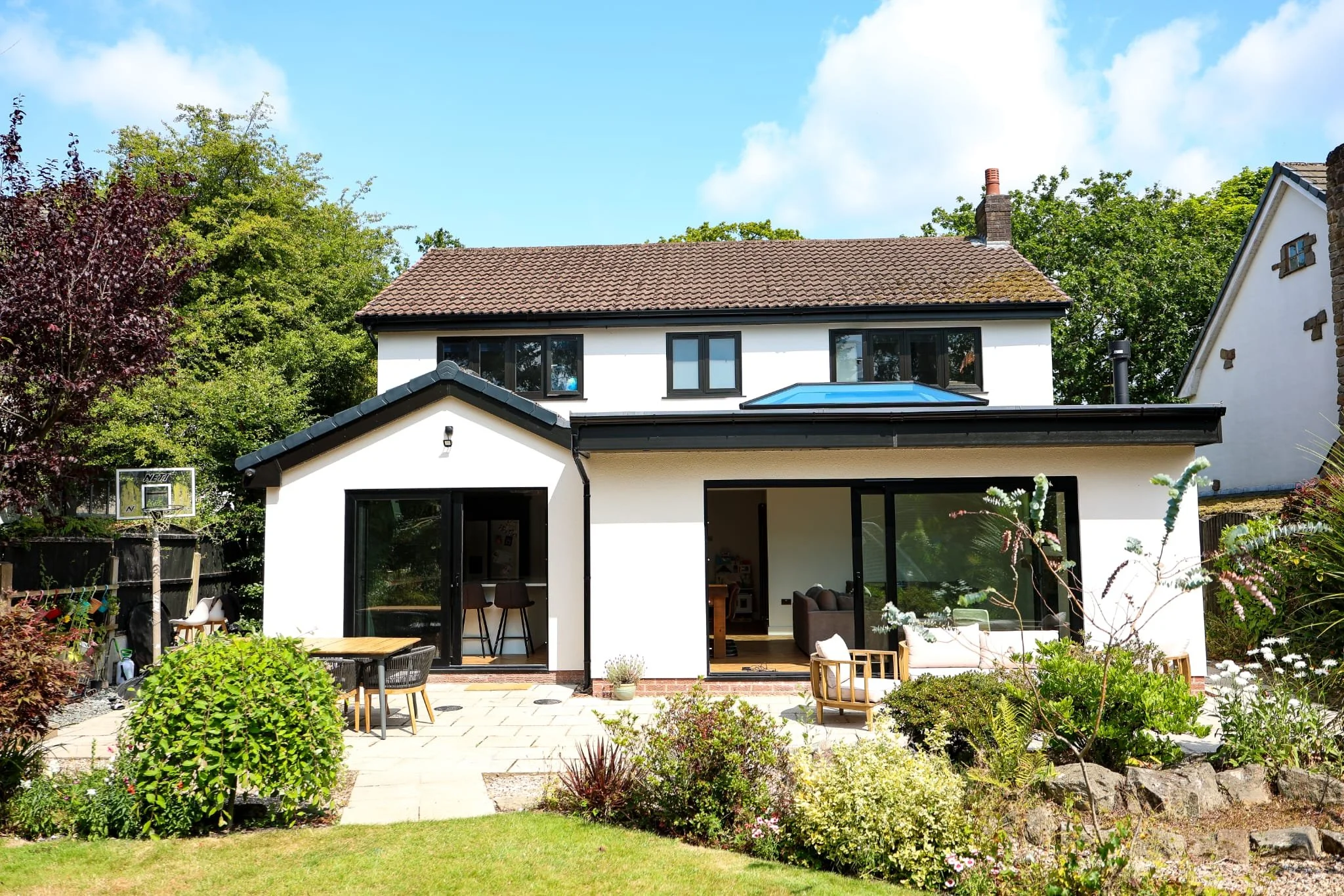 Backyard view of a modern white house with black window frames and a brown tiled roof, surrounded by green trees and a landscaped garden with outdoor furniture.