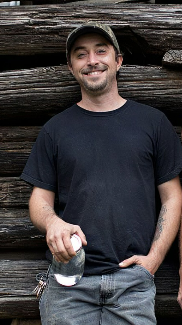 A man smiling, wearing a black t-shirt and camo hat, standing in front of a large wooden log wall, holding a drink and keys.