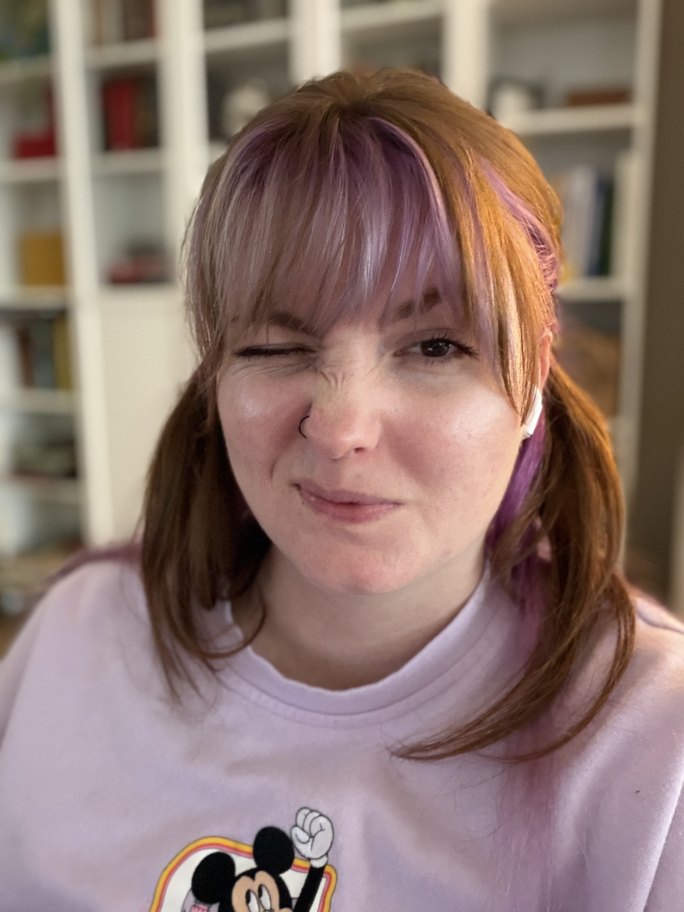 A woman with purple and red hair wearing a light purple sweater squints mischievously at the camera