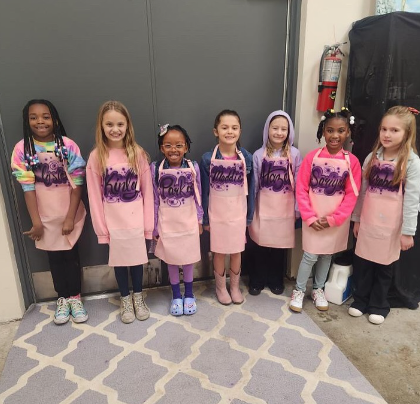 Seven smiling young girls standing side by side, wearing pink aprons with their names written in purple, in an indoor setting with a gray door and a fire extinguisher in the background.