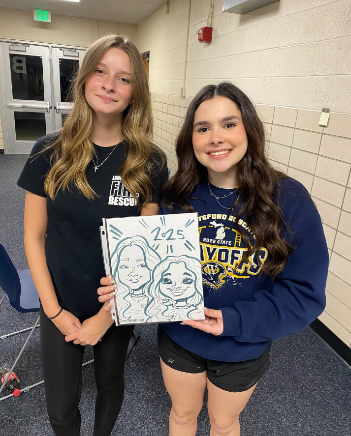 Two young women standing inside a school hallway, smiling at the camera, one holding a drawing of two women with '2025' written above, and the other wearing a 'Bayford' and '2022 State' sweatshirt.